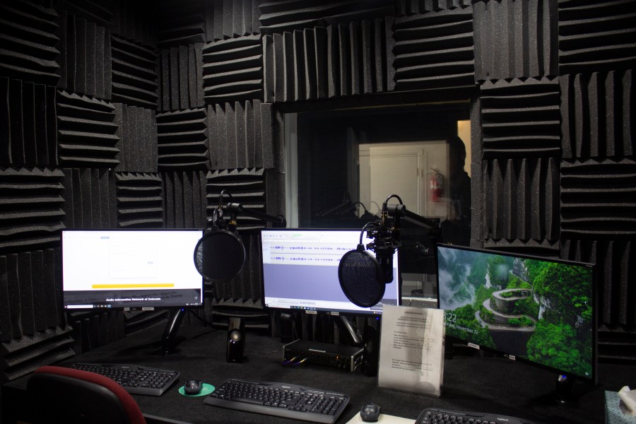 a photo of a studio covered in acoustic foam. 3 computers show audio software, and 2 mics hover above the desk.